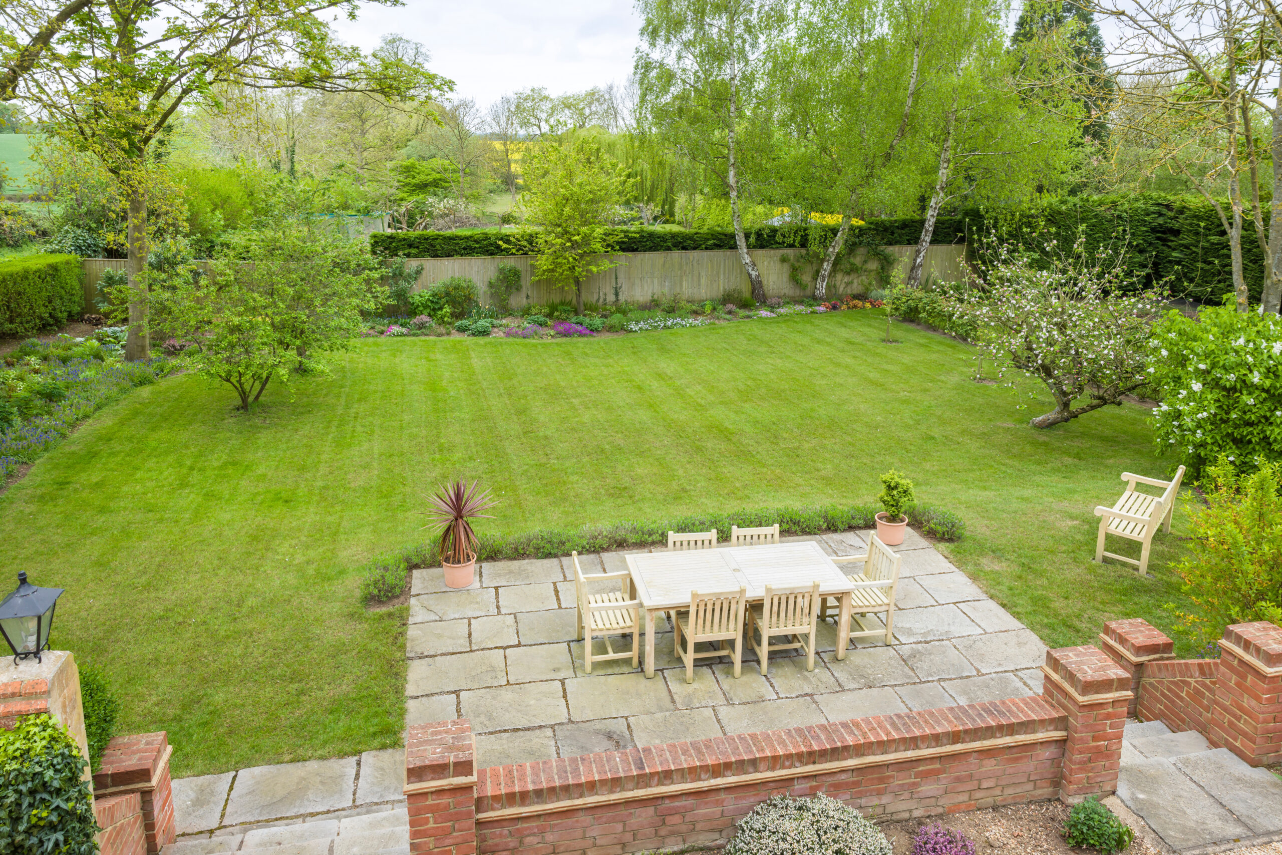 A neatly mowed backyard features a stylish timber decking area with a wooden dining table and chairs, garden bench, and lush green trees and plants surrounding a large lawn—perfectly maintained by a skilled landscaper.